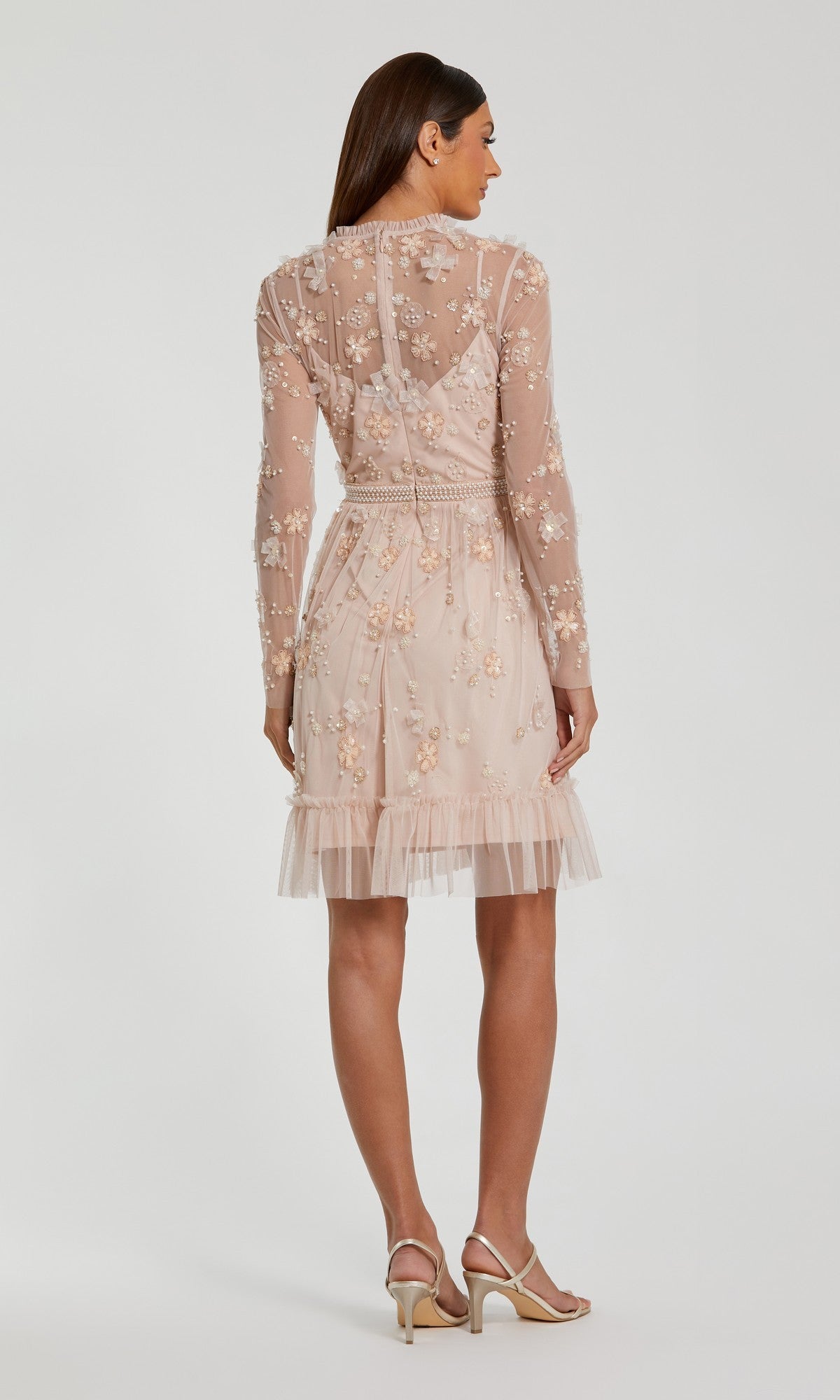 A woman stands facing away, wearing the Mac Duggal 9140 short black wedding guest dress with long sleeves and a ruffled hem. She has straight brown hair and wears beige heeled sandals.